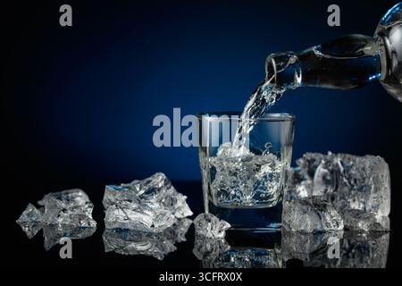 Kristallklarer Wodka oder Gin wird über Eis in ein Glas gegossen. Wodka und Eisstücke auf schwarzem reflektierendem Hintergrund. Kopierbereich. Stockfoto