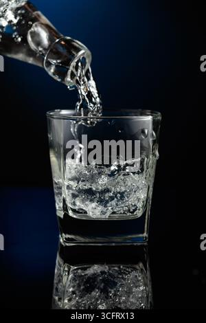 Kristallklarer Wodka oder Gin wird über Eis in ein Glas gegossen. Wodka mit Eis auf schwarzem reflektierendem Hintergrund. Stockfoto