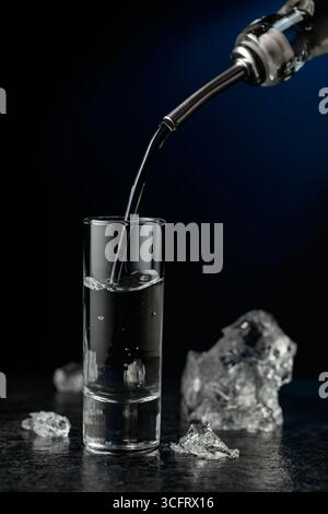 Kristallklarer Wodka oder Gin wird in ein Glas gegossen. Transparentes alkoholisches Getränk mit Eisstücken auf schwarzem Hintergrund. Stockfoto