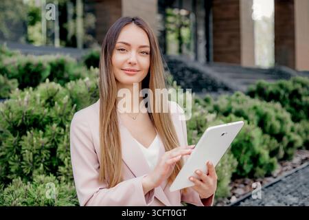 Eine junge kaukasische Frau in einem Business-Anzug hält ein Tablet in der Hand und schaut auf die Kamera vor dem Hintergrund eines Bürogebäudes mit Stockfoto