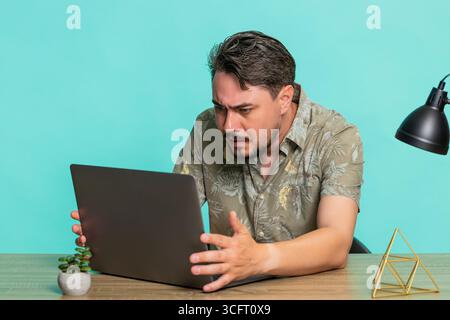 Ein Mann mittleren Alters sitzt am Tisch und erhält schlechte Nachrichten. Er erlebt eine Niederlage bei Online-Spielen auf einem Laptop und zeigt eine deutliche Enttäuschung. Junger Erwachsener, isoliert auf blauem Hintergrund, der sich verärgert fühlt Stockfoto