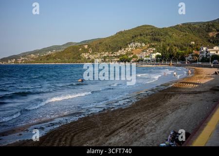 Blick auf die Sandküste und Wellen entlang der Küste von Karamürsel, Kocaeli, Türkiye, mit grünen Hügeln und Häusern im Hintergrund. Ein paar Leute sind es Stockfoto