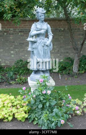 Eine anmutig detailreiche Statue zeigt eine Frau in historischer Kleidung, die tagsüber in einem ruhigen Garten inmitten lebendiger Blumen steht. Stockfoto