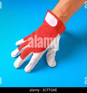 Dieses eindrucksvolle Bild zeigt die Hand eines Mannes in einem hellroten Arbeitshandschuh, der sich wunderbar mit einem satten blauen Hintergrund abhebt. Die lebendige Farbe Stockfoto