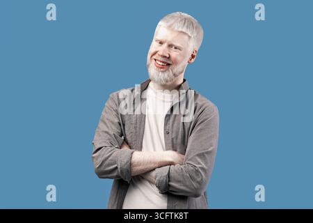 Selbstbewusste Person. Lächelnder Albino-Typ mit gefalteten Armen, der in die Kamera schaut, isoliert über blauem Studiohintergrund posiert Stockfoto
