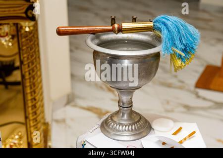 Nahaufnahme eines Weihwasserbehälters aus Metall mit einem traditionellen Aspergillum (Pinsel), der zum Besprühen von Weihwasser während der religiösen Zeremonie verwendet wird Stockfoto
