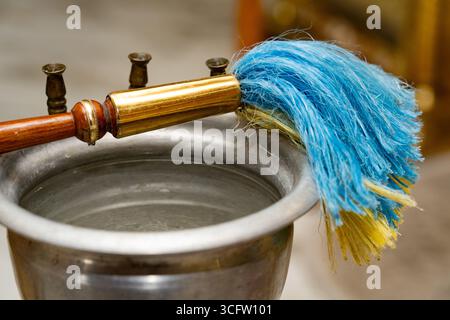 Nahaufnahme eines Metall-Weihwasserbehälters mit einem traditionellen Aspergillum, das zum Besprühen von Weihwasser während religiöser Zeremonien verwendet wird Stockfoto