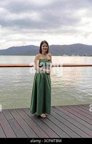 Eine elegante Frau in einem grünen Kleid, die auf einer Holzpromenade vor einem malerischen Küstengrund mit Bergen und bewölktem Himmel steht Stockfoto