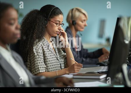 Junge Erwachsene Frau, die ein Headset trägt, arbeitet am Computer in modernen Büros, sitzt am Schreibtisch mit Kollegen, einschließlich einer Frau mittleren Alters, die ein Headset im Hintergrund verwendet Stockfoto