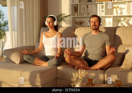 Ein paar Amateur-Yogis, die zu Hause Yoga machen, hören Online-Guide von Kopfhörern, die bei Sonnenuntergang auf einem Sofa sitzen Stockfoto