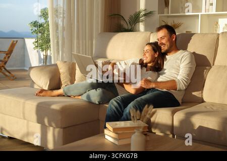 Glückliches Paar bei Sonnenuntergang mit Laptop auf einer Couch sitzend und zu Hause umarmt Stockfoto