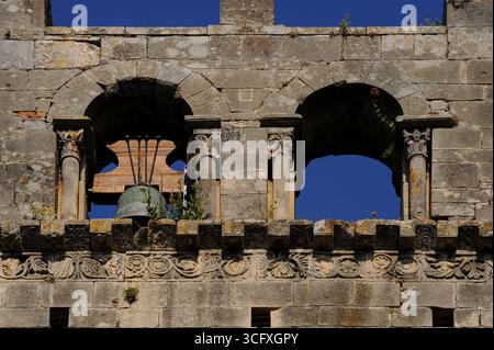 Ein „grüner Mann“, dessen Laub aus dem Mund wächst, ziert die Hauptstadt einer Säule, die den runden romanischen rechten Bogen des Glockenturms über dem Turm der Kathedrale unserer Lieben Frau von Nazareth in Vaison-la-Romaine, Vaucluse, Provence-Alpes-Côte d’Azur, Frankreich, stützt. Eine große Bronzeglocke hängt im linken Bogen, während ein mondähnliches Gesicht aus dem Blumenfries darunter blickt. Stockfoto