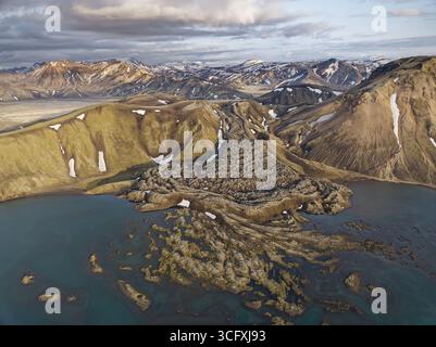 Blick aus der Vogelperspektive auf zerklüftete, moosbedeckte Hügel, die in das ruhige Seenwasser unter der Kulisse schneebedeckter Gipfel fallen, die Highlands, Island. Stockfoto