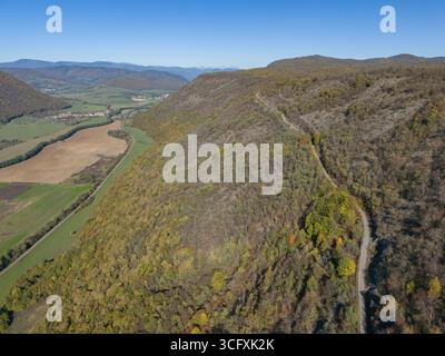Aus der Vogelperspektive schlängelt sich eine gewundene Straße durch einen bewaldeten Hügel, im Kontrast zu den offenen Feldern und entfernten Bergen, Plešivec, Region Košice, Slowakei. Stockfoto