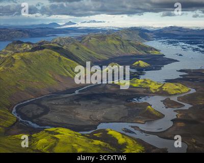 Blick aus der Vogelperspektive auf das grüne Hochland, das auf die geflochtenen Kanäle des Gletscherflusses trifft, im Kontrast zu dunklem vulkanischem Sand unter einem riesigen isländischen Himmel, Hochland, Island. Stockfoto