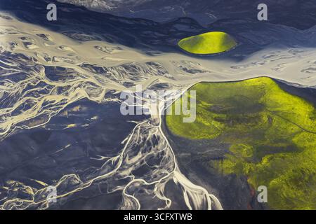 Aus der Vogelperspektive sehen Sie geflochtene Flüsse, die sich durch dunkle vulkanische Ebenen schlängeln, im Kontrast zu lebendigen grünen Vegetationsflächen, die eine surreale Landschaft erschaffen, Island. Stockfoto