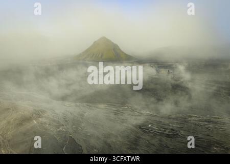 Majestätisch erhebt sich der Blick auf einen einsamen Berg, der von ätherischem Nebel umhüllt ist, aus der dunklen, strukturierten Vulkanlandschaft im Hochland, Island. Stockfoto