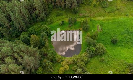 Aus der Vogelperspektive auf einen kleinen Waldteich mit Wolken, die sich auf der Wasseroberfläche spiegeln, umgeben von üppigem Grün und Bäumen im ländlichen Litauen Stockfoto