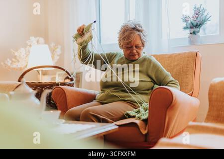 Seniorinnen stricken in gemütlicher und gut beleuchteter Inneneinrichtung auf einem bequemen Sessel, umgeben von einer ruhigen Einrichtung. Stockfoto