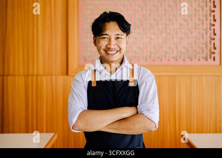 Porträt eines lächelnden asiatischen Küchenchefs mit schwarzer Schürze und weißem Hemd, der mit überkreuzten Armen in einem modernen Holzrestaurant steht. Ideal Stockfoto