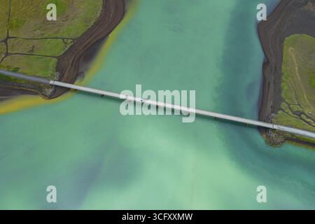 Aus der Vogelperspektive einer Betonbrücke, die durch das türkisfarbene Wasser schneidet, im Kontrast zur dunklen, strukturierten Erde auf beiden Seiten, isländischen Straßen, Island. Stockfoto