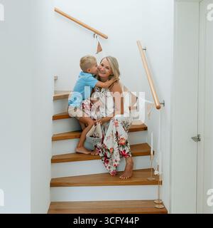 Mutter sitzt auf Holztreppen, während ihr Kind sie liebevoll umarmt. Die Szene fängt einen zarten Moment der Nähe und des Glücks ein, mit Wärme Stockfoto