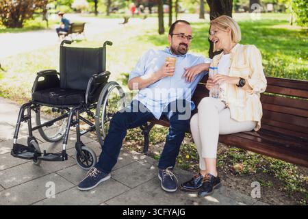 Ein Mann im Rollstuhl verbringt Zeit mit seiner Mutter im Park. Querschnittsgelähmter spricht mit der Frau. Stockfoto