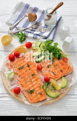 Gegrilltes Lachsfilet auf einem runden Holzbrett mit Zitrone, Avocado, Tomaten, Zwiebeln und Petersilie auf weißem Holztisch mit Gabeln und Serviette, vert Stockfoto