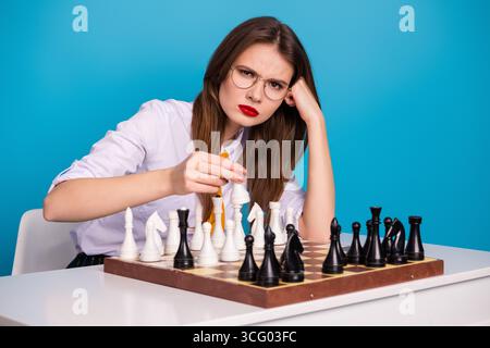 Müde Frau, die Schachfiguren auf einem Spielbrett bewegt, während sie auf einem blauen Hintergrund sitzt Stockfoto