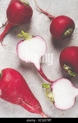 Leuchtend rote Radieschen sind auf einer Theke angeordnet, wobei ein Rettich in der Mitte geschnitten wird, um das klare weiße Innere freizulegen. Die grünen Oberteile sorgen für einen Hauch von Frische Stockfoto
