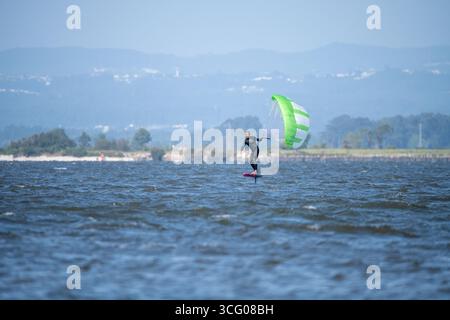 Ein Mann mittleren Alters in einem Taucheranzug gleitet auf einem Tragflächenbrett mit einem grünen und weißen Drachen, mit Hügeln und einer Küstenlinie in der Ferne Stockfoto