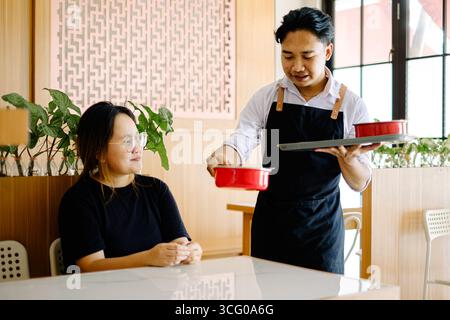 Ein lächelnder asiatischer Kellner auf der Schürze serviert einer sitzenden Frau in einem hellen, modernen chinesischen Restaurant Essen in roten Schüsseln Stockfoto