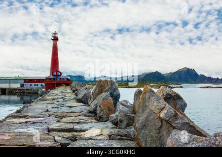 Der Leuchtturm Red Andenes steht hoch auf einem felsigen Pier mit ruhigem Wasser und fernen Bergen unter einem bewölkten Himmel und zeigt die Schönheit der Küstenlandschaft und die maritime Schönheit Stockfoto