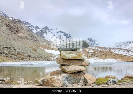 Ein ausgeglichener Steinhaufen steht an einem ruhigen, teilweise gefrorenen Bergsee, der von schneebedeckten Gipfeln eingerahmt wird Stockfoto