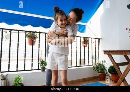 Ein zärtlicher Moment zwischen einer Mutter, die ihre Tochter auf einer gemütlichen Terrasse mit Pflanzen umarmte. Sonnenlicht verstärkt die fröhliche Bindung, die sie an dieser Seite teilen Stockfoto