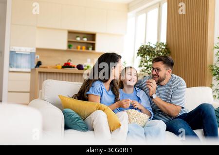 Lachen erfüllt die Luft, während eine Familie gemeinsam Popcorn genießt und fröhliche Momente in ihrem stilvollen Wohnzimmer während eines entspannten Abends teilt. Stockfoto