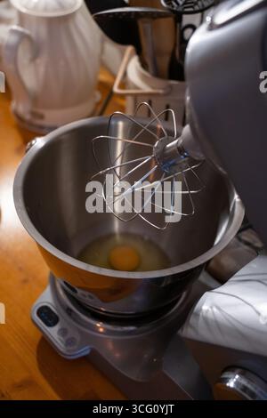 Frisches Ei in einer Edelstahlrührschüssel eines elektrischen Standmixers, bereit zum Rühren auf einer hölzernen Küchentheke Stockfoto
