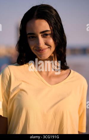 Junge Frau lächelt am Strand bei Sonnenuntergang und trägt ein gelbes T-Shirt Stockfoto