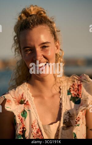 Porträt einer lächelnden jungen Frau mit lockigen blonden Haaren zur goldenen Stunde Stockfoto