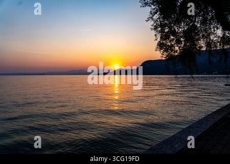 Sonnenuntergang Reflektionen über dem Lago di Gardasee mit Silhouettenhügeln und Baum, Italien Stockfoto