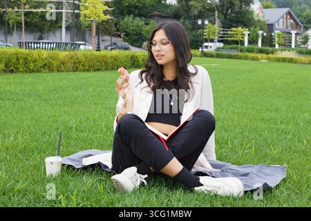 Junge indische College-Studentin sitzt auf Gras mit Buch, Donut und Getränk, genießt Outdoor-Studium auf einer Decke, Symbol für moderne Bildung, ausgewogenes li Stockfoto