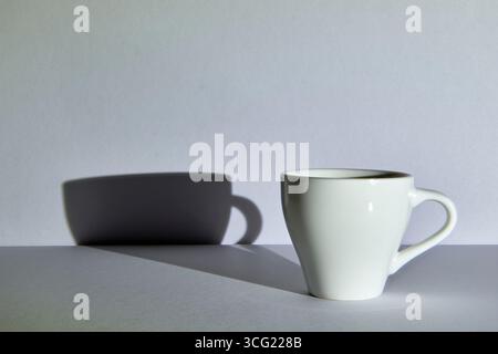 Ein einzelner weißer Becher wirft einen starken Schatten auf einem grauen Hintergrund. Ein minimalistisches Stillleben mit hohem Kontrast, das Einfachheit, Dualität oder den Anfang repräsentiert Stockfoto