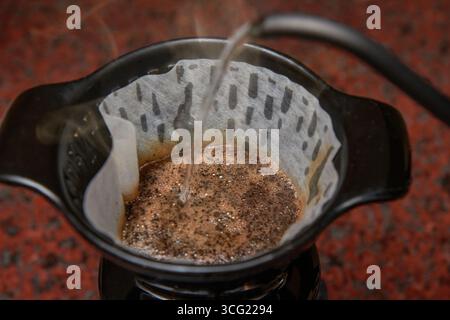 Gießen Sie heißes Wasser aus einem Schwanenhals-Wasserkocher in einen Filter mit gemahlenem Kaffee, während Dampf steigt. Ein dynamischer Schuss der Zubereitungsmethode Capturi Stockfoto