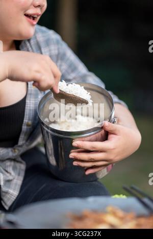 Eine übergewichtige indonesische südostasiatische Frau, die beim Camping einen Metallbehälter mit gekochtem Reis hält. Die Szene des Lebensstils des Genießens von Fo Stockfoto