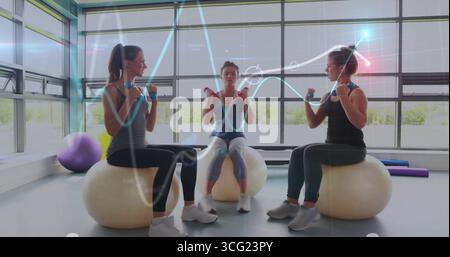 Frauen in Sportbekleidung, die Kurzhanteln auf Trainingsbällen im Fitnessstudio mit großen Fenstern lockern Stockfoto