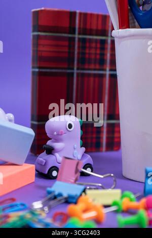 Verspielter Miniatur-Dinosaurier, der ein violettes Spielzeugauto mit farbenfrohen Schreibwaren, Radiergummis, Büroklammern und Stecknadeln vor einem leuchtend violetten Hintergrund fährt Stockfoto