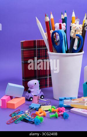 Schreibwaren wie Buntstifte, Scheren, Radierer und Clips vor violettem Hintergrund sorgen für eine lebendige Schulszene Stockfoto