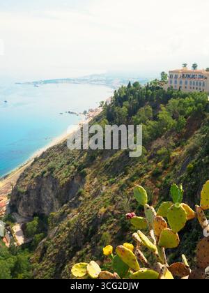 Blick auf Taormina an den Klippen mit mediterraner Küste, Kaktuspflanzen im Vordergrund und Küstenlandschaft entlang des Ionischen Meeres in Sizilien Stockfoto