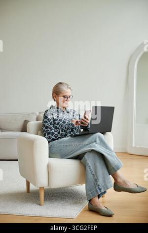 Ältere kaukasische Frau, die ihr Smartphone durchsucht, während sie drinnen an einem Laptop arbeitet Stockfoto