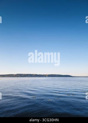 See am Morgen. Kamera nahe der ruhigen Wasseroberfläche des großen Sees kurz nach Sonnenaufgang. Steigungshimmel über dem Montanufer des Flusses. Stockfoto